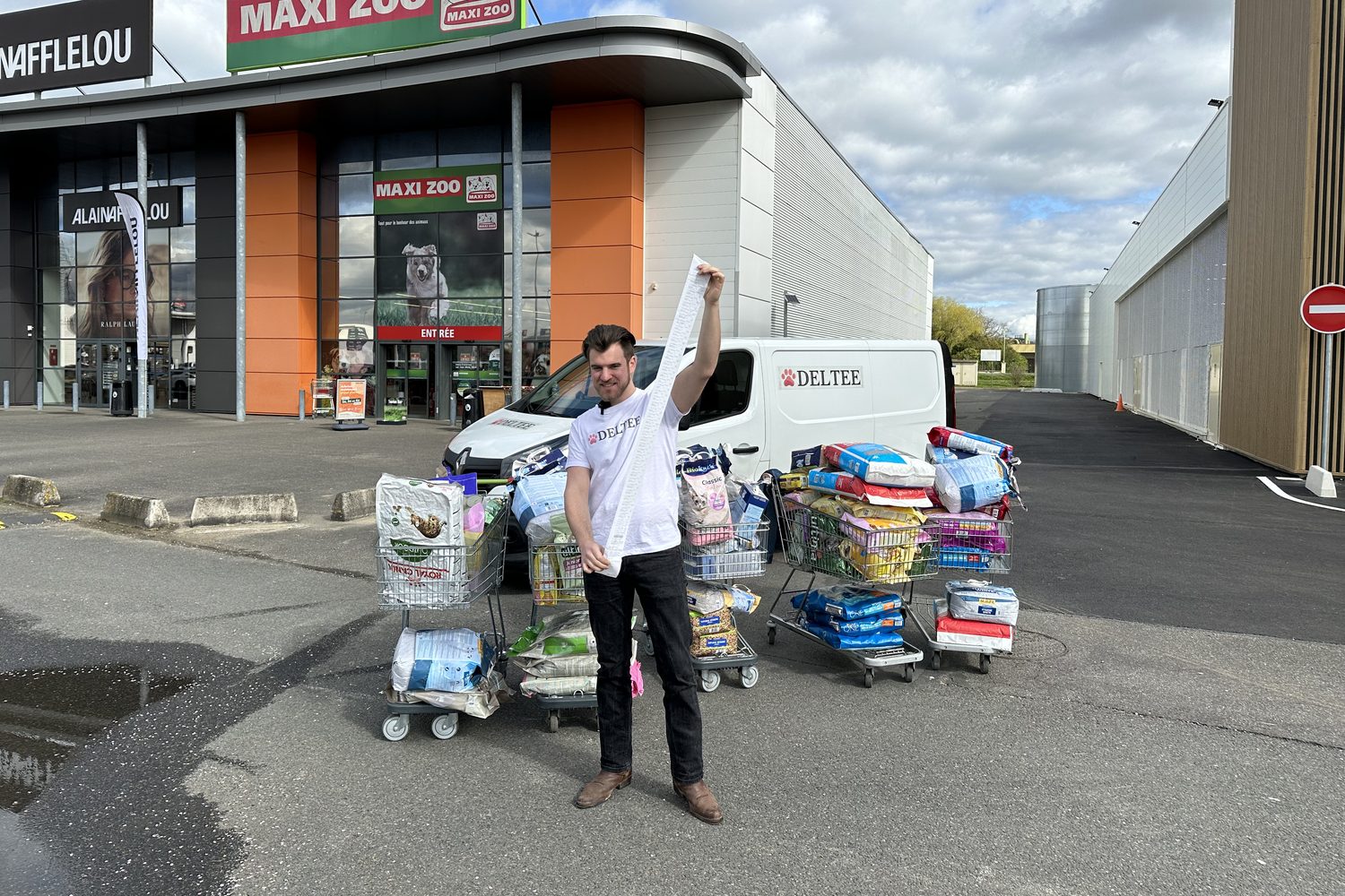 Un membre de l'équipe Deltee, bras levé, devant des chariots remplis de croquettes et de litière pour les refuges